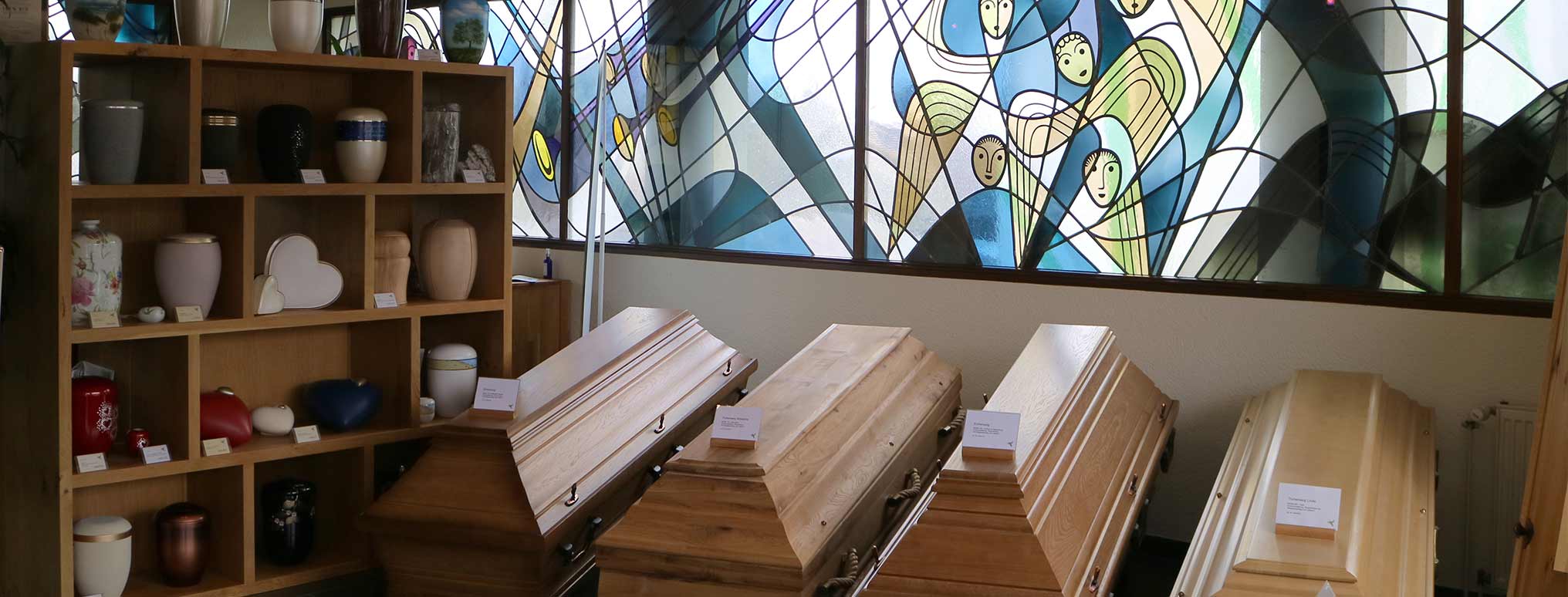Ausstellungsraum mit einer Auswahl an Holzsärgen und Urnen, stilvoll präsentiert vor einem kunstvollen Buntglasfenster mit Engelmotiven.